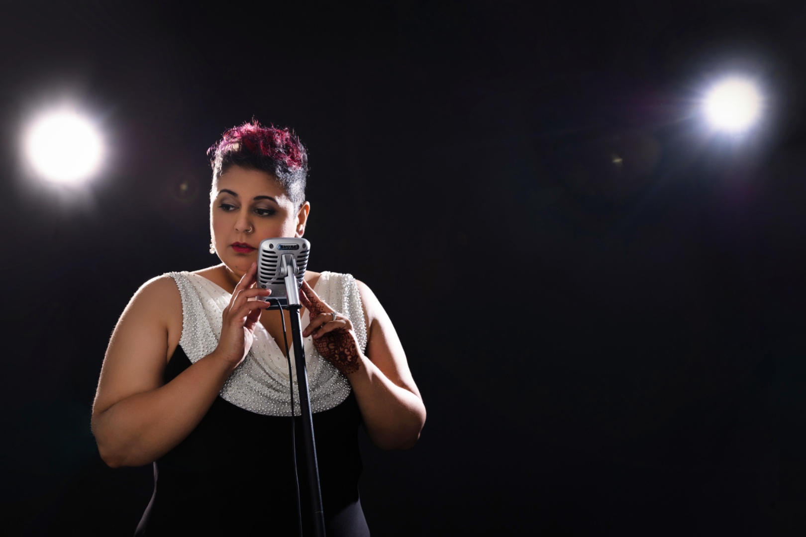Farah Kidwai performing at a vintage microphone under dramatic stage spotlights, wearing a black gown with a beaded white drape
